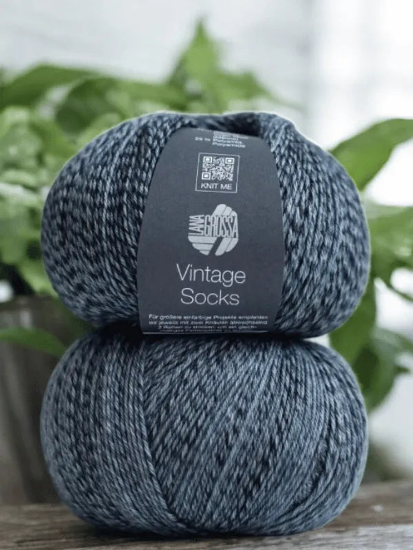 Two skeins of Lauri Fingerin sukkaohje + Lana Grossa Vintage socks yarn in dark gray and white are stacked with soft green plants blurred in the background—ideal for your next villasukat project.