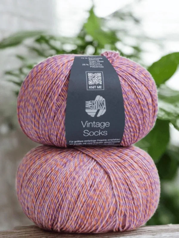 Two balls of Lana Grossa Vintage Socks yarn in purple, pink, and yellow are stacked vertically. Soft green leaves blur behind—ideal for knitting villasukat or using the Lauri Fingerin sukkaohje pattern.