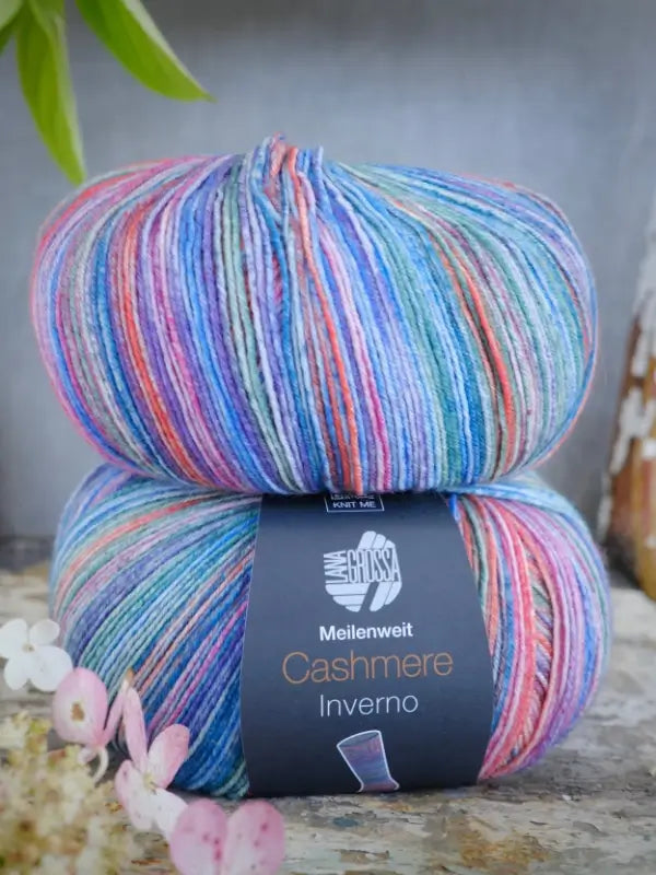 Two skeins of multicolored yarn, featuring shades of blue, purple, pink, and green, are stacked. The label reads Lana Grossa Meilenweit 100 Cashmere Inverno—a luxurious cashmere sock yarn ideal for your most exquisite knitting projects. Small flowers rest in the foreground.