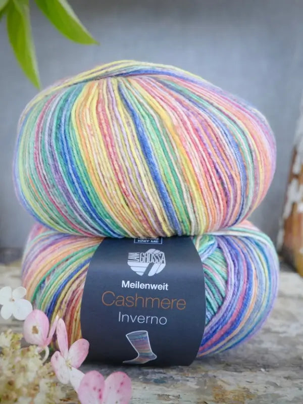Two skeins of rainbow-striped Lana Grossa Meilenweit 100 Cashmere Inverno yarn are stacked on a rustic surface with delicate pale pink flowers in the foreground, showcasing the luxurious softness of this cashmere sock yarn.