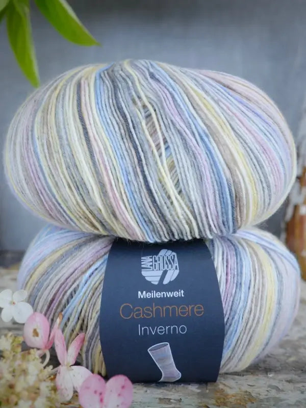 A skein of Lana Grossa Meilenweit 100 Cashmere Inverno yarn in pastel multicolors rests on a surface adorned with delicate flowers, its luxurious softness and quality evocative of fine Kashmir sukkalanka, all set against a blurred background.