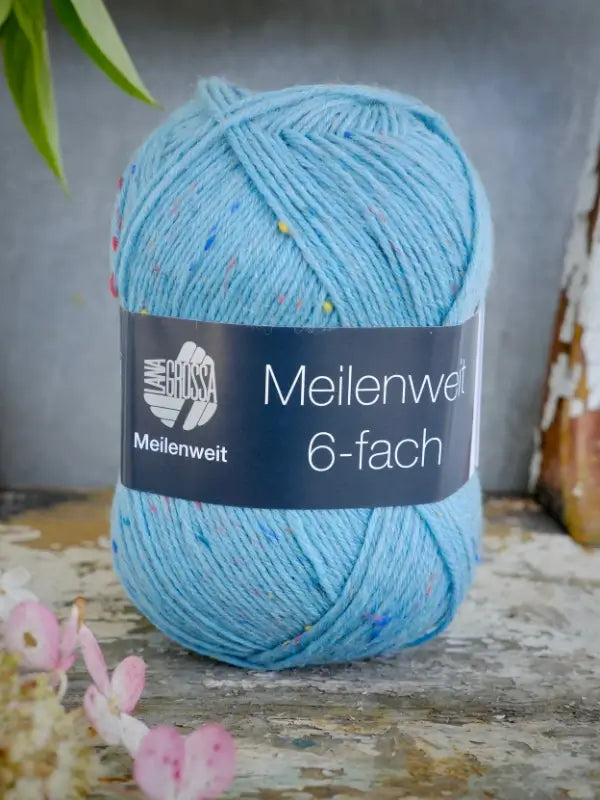 A ball of Lana Grossa Meilenweit 6 Tweed yarn in light blue with colorful specks rests on a rustic surface, accompanied by a plant and pink flowers in the background. This charming setting is perfect inspiration for starting your next neulesetti or for giving the Lauri sukkaohje sport + Lana Grossa Meilenweit 6 Tweed pattern a try.