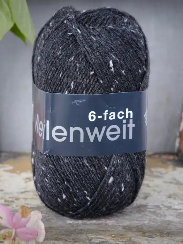 A skein of Lana Grossa Meilenweit 6 Tweed yarn in dark gray with tweed speckles rests on a mottled surface, with a pink flower partially visible in the lower left corner.