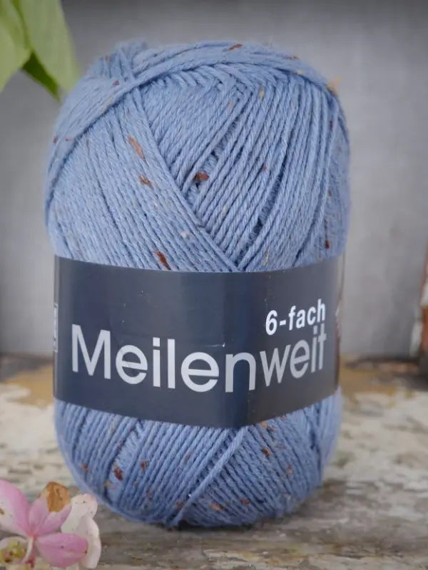 A skein of Lana Grossa Meilenweit 6 Tweed yarn in blue with delicate brown speckles rests on a rustic surface, accented by a hint of pink flower in the foreground. This six-ply (kuusisäikeinen) tweed sock yarn provides a classic texture, making it ideal for cozy projects that need understated detail.