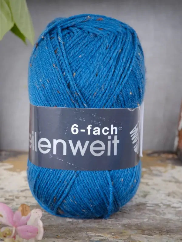 A skein of Lana Grossa Meilenweit 6 Tweed yarn, featuring blue tones with flecks and marked 6-fach, rests on a weathered surface. The label highlights its quality as a tweed-sukkalanka. A pink flower appears in the lower left corner.