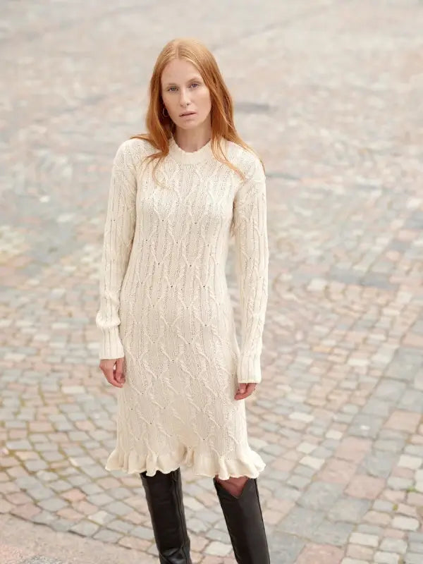 A woman with long red hair stands on a stone-paved street, embodying Nordic Chic in a cream-colored, long-sleeve knit dress with a ruffled hem and black knee-high boots, capturing the timeless style celebrated in “Leeni Hoimela – Skandityyli, Ajattomia ohjeita nykyneulojalle.”.
