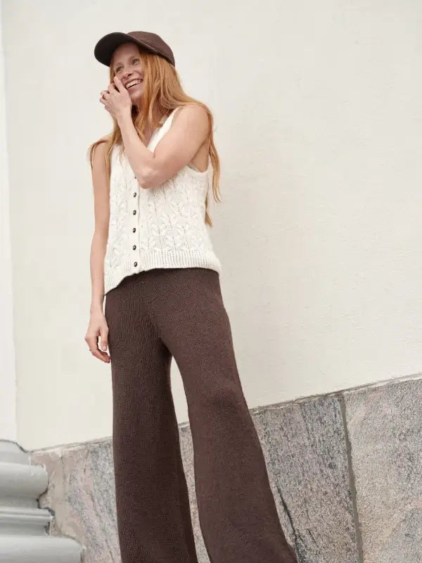 A woman exemplifying Nordic Chic stands on stone steps, smiling with one hand near her face against a light-colored wall, dressed in a white knit sleeveless top and brown wide-leg pants—capturing the timeless elegance featured in *Leeni Hoimela – Skandityyli, Ajattomia ohjeita nykyneulojalle*.