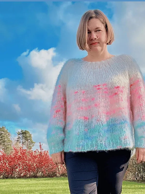 A person with short blonde hair stands on grass outdoors, wearing an oversized pastel sweater in pink, blue, and white—knit from the Lempi pusero neuleohje pattern. The bright sky features puffy clouds, with trees and red bushes behind them.