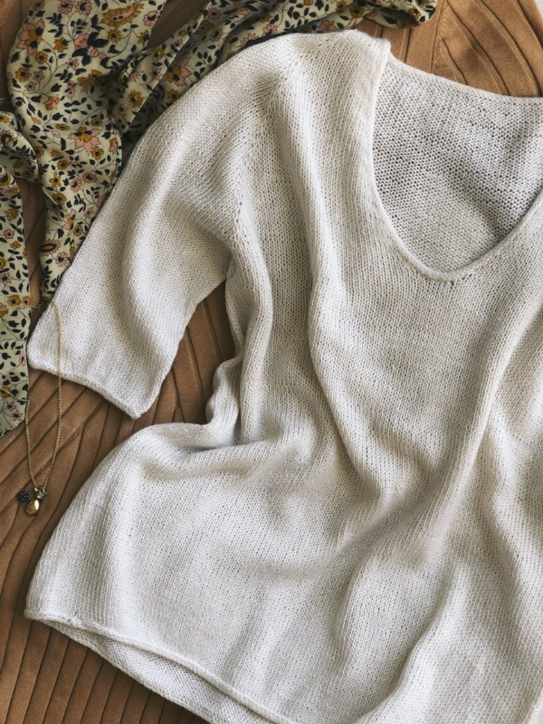 The PetiteKnit - Cumulus Tee neuleohje is a lightweight, sheer white knit tee with a wide V-neck, displayed flat on a ribbed beige surface beside a floral scarf and a delicate gold charm necklace.