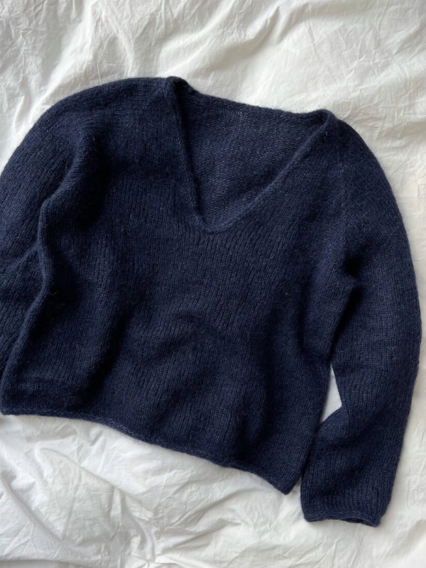 A dark navy blue, long-sleeve knit sweater with a deep V-neck is laid flat on a light fabric background, inspired by the PetiteKnit - Cumulus Blouse neuleohje design.