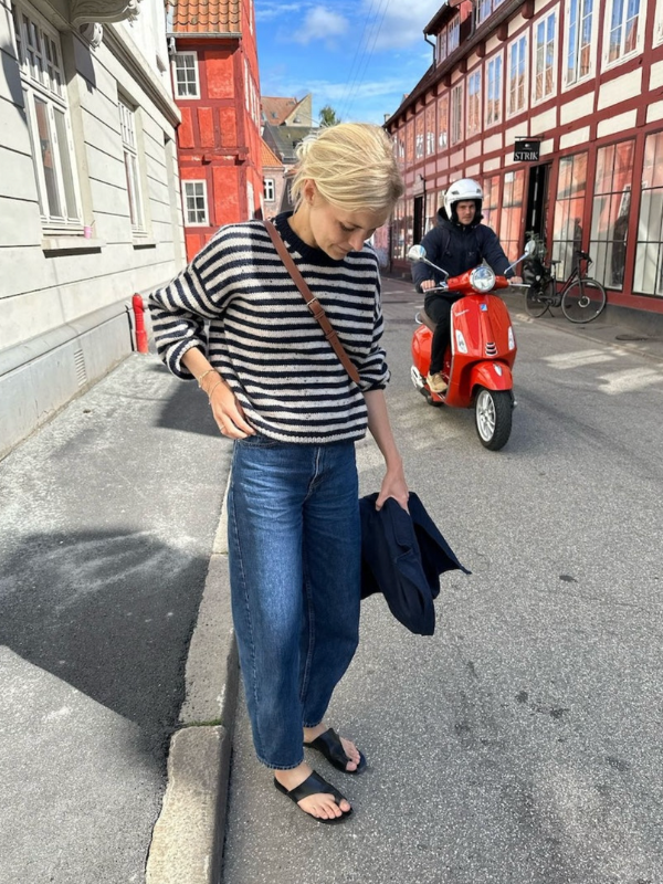 A woman in a PetiteKnit Olga sweater neuleoohje, blue jeans, and sandals stands on a street lined with colorful buildings. A red scooter with a helmeted rider passes by behind her as she looks down.