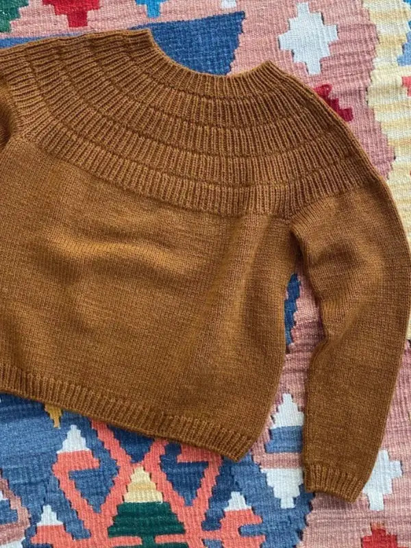 The PetieKnit Anker’s Sweater - My Size neuleohje - Suomi, a brown knitted sweater featuring ribbed detailing on the yoke and cuffs inspired by the classic Anker’s Sweater, is laid flat on a colorful geometric-patterned rug.