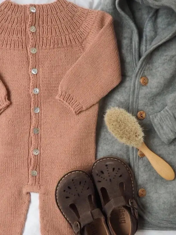 A pink knit baby romper with buttons, inspired by the delightful PetitKnit Anker’s Onesie Neuleohje – Suomi, a gray hooded sweater, a wooden brush with natural bristles, and a pair of brown toddler sandals are arranged on a light surface.