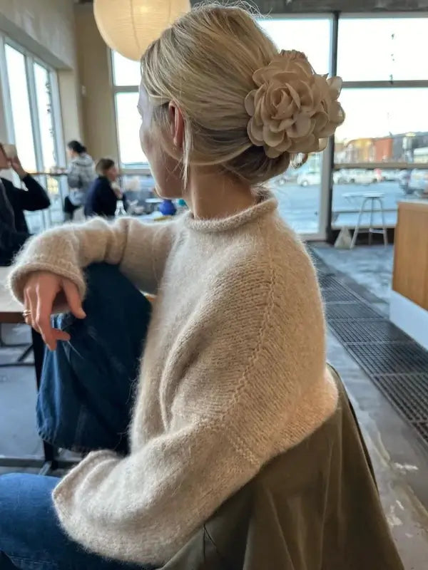 A woman with blonde hair styled in a bun and adorned with a large beige flower sits in a modern café, gazing thoughtfully out the large windows. She is wearing a cozy sweater knitted from the PetiteKnit -Cloud Sweater Neuleohje pattern in soft beige, paired with blue jeans, while her coat rests casually over her arm. The inviting atmosphere of the café provides the perfect spot to relax with her coffee and flip through the latest neuleohje magazine for inspiration.