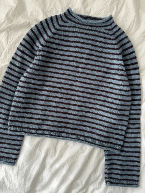 The PetiteKnit Frankie sweater neuleohje suomi, a knitted long-sleeve sweater with blue and dark brown horizontal stripes, is laid flat on a white, slightly wrinkled fabric background.