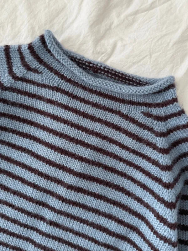 A close-up of the PetiteKnit Frankie sweater neuleohje suomi, featuring horizontal light blue and dark brown stripes with raglanlisäykset, lying on a white textured fabric background.