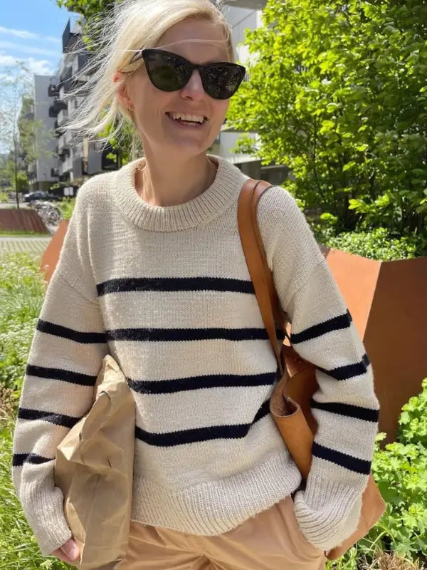 A smiling person wearing sunglasses and the PetiteKnit Marseille Sweater neuleohje- Suomi in cream with black stripes, carrying a brown bag and a paper package, stands outdoors on a sunny day with greenery and buildings in the background.