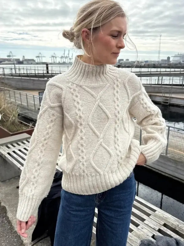 A woman wearing a cream PetiteKnit Saga sweater neuleohje Suomi and blue jeans stands outdoors near a waterfront, with docks and cranes visible in the background.