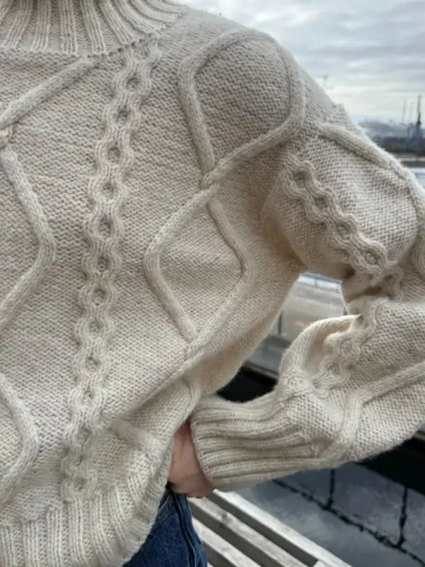 A close-up of a person wearing a cream cable knit sweater with diamond and rope patterns, made using the PetiteKnit Saga sweater neuleohje Suomi, standing outdoors near a railing with a cloudy sky in the background.