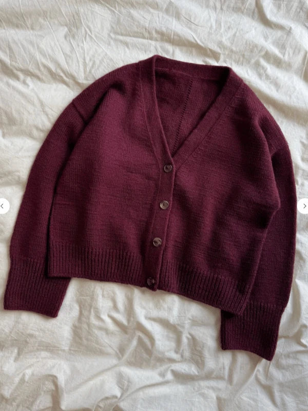 The PetiteKnit Scarlet Cardigan -ENGLISH is a deep burgundy knit with a classic English pattern, featuring long sleeves, ribbed cuffs and hem, and three dark buttons down the front, displayed flat on a light wrinkled fabric background.