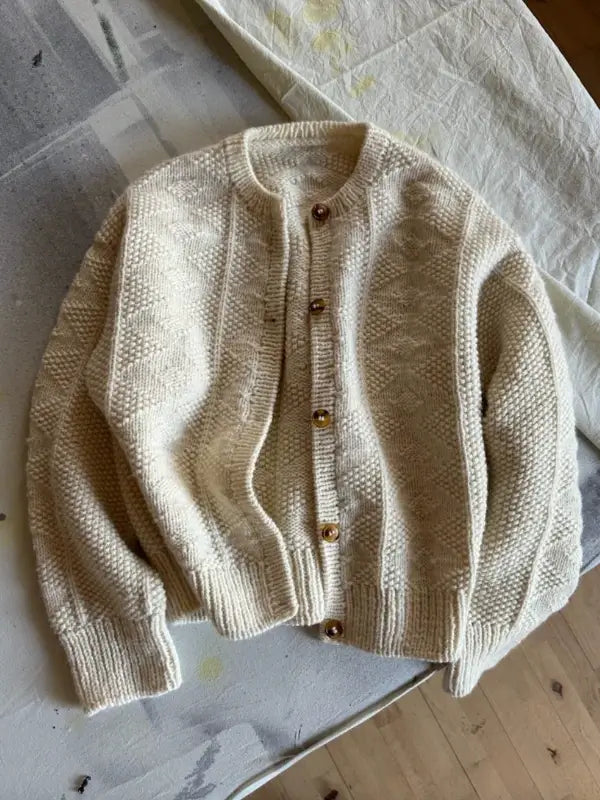 The PetiteKnit - Esther Jacket Neuleohje is a cream-colored cardigan featuring a textured knit pattern, wooden buttons, and ribbed cuffs and hem, displayed flat on a light cloth.