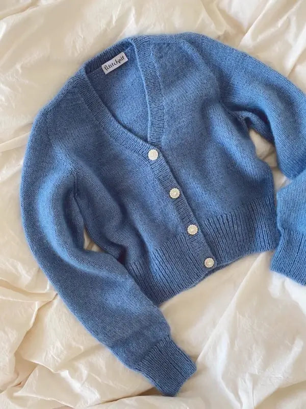 The PetiteKnit - April Cardigan neuleohje is a blue, long-sleeved knit sweater with a V-neckline, white buttons, and ribbed hem, cuffs, and button band. Displayed flat on a cream fabric background.