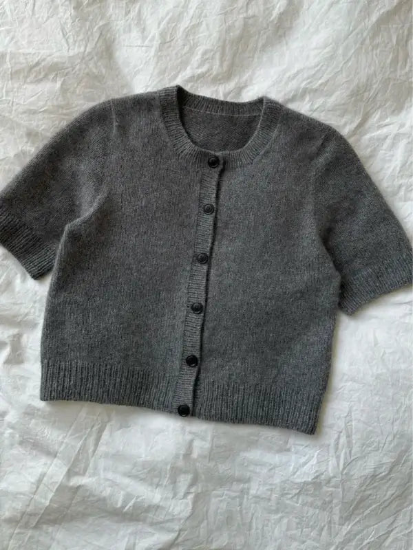A PetiteKnit Maggie Cardigan neuleohje in grey, short-sleeved and button-up, is laid flat on a white, textured fabric background.
