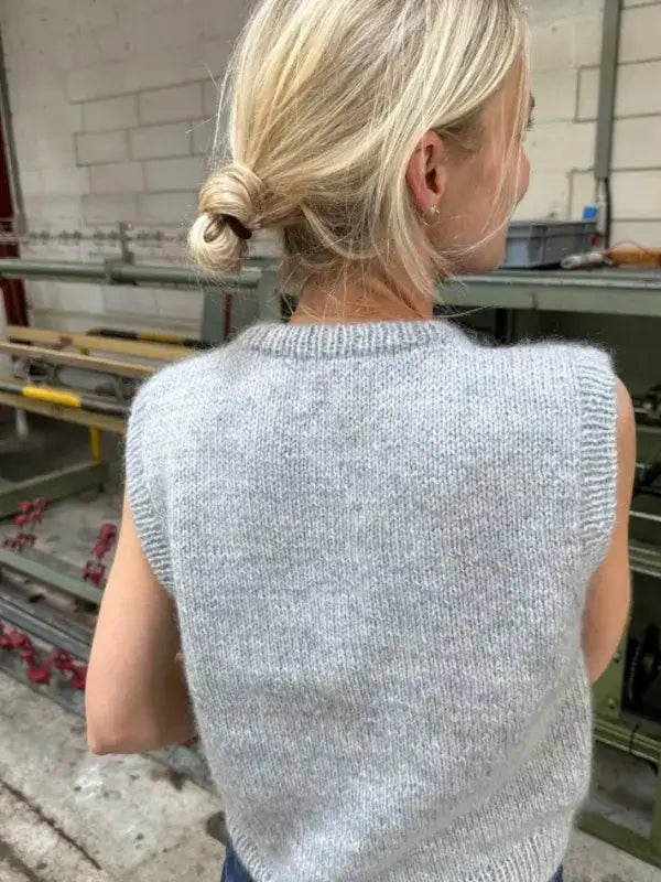 A person with blonde hair in a low bun is wearing a sleeveless gray knit PetiteKnit Novice Slipover neuleohje, standing indoors near industrial shelves and a brick wall.