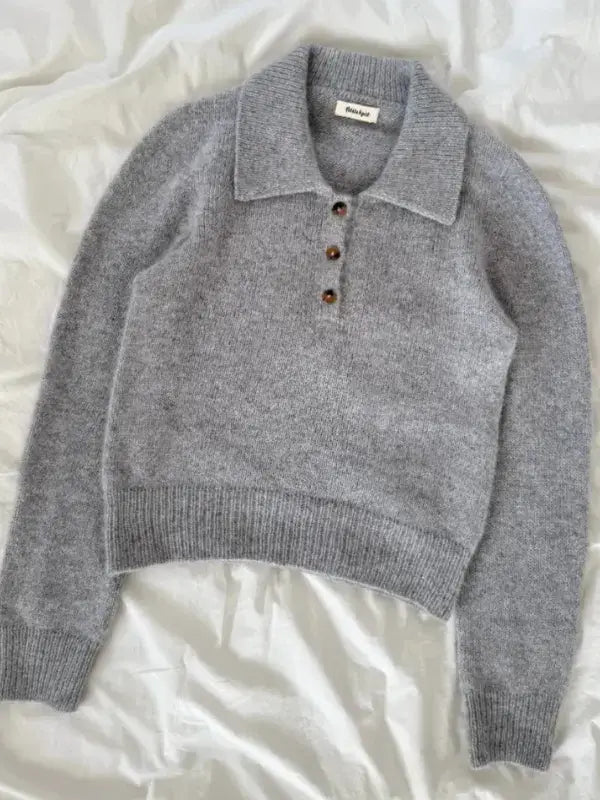 A gray knitted sweater with a wide collar and four brown buttons on the front, laid flat on a white, slightly wrinkled surface. This cozy piece is made following the PetiteKnit Petra Polo Neuleohje pattern and includes a suomenkielinen printti for easy knitting guidance.