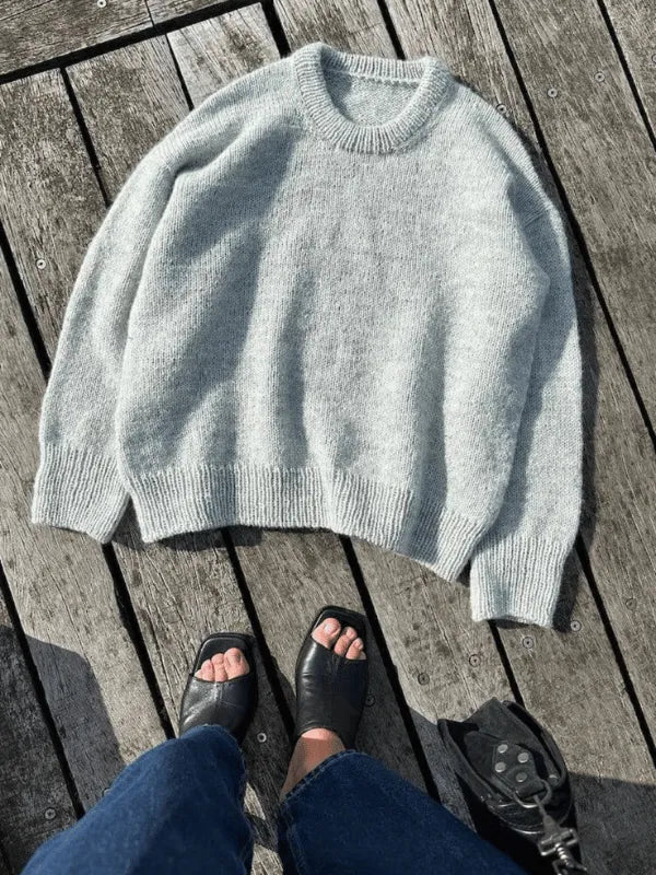 The PetiteKnit Sonja Sweater neuleohje Suomi, a light gray top-down knit, is shown laid flat on wood. Blue jeans and black open-toe shoes are partly visible with a corner of a black bag. Neuleohje saatavilla.