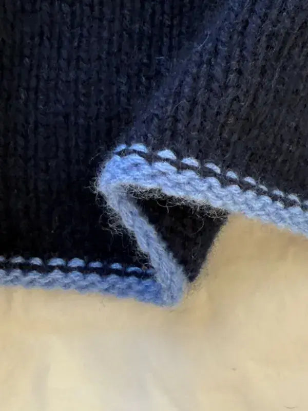 A close-up of dark blue knitted fabric with a light blue border, showing the edge of the PetiteKnit Uma Scarf neuleohje folded over on a light surface.