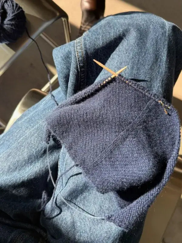 A close-up of blue knitting in progress on wooden needles, resting on someone’s lap clad in jeans. Sunlight casts gentle shadows across the scene as soft cashmere yarn transforms into what will become the cozy PetiteKnit Uma Scarf neuleohje.