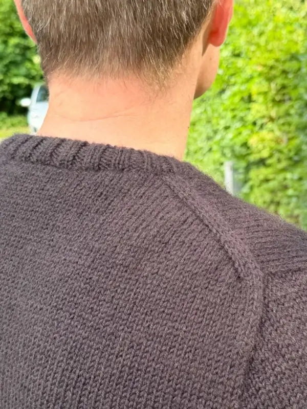 A close-up of the back of a person wearing a dark Petiteknit Niels Sweater neuleohje, Suomi, showing the collar and shoulder seam, with short light brown hair and greenery in the background.