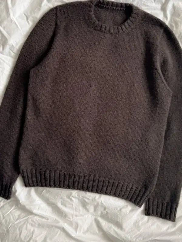 A dark brown, long-sleeved knitted sweater—Petiteknit Niels Sweater neuleohje, Suomi—is laid flat on a light, crinkled fabric background. The sweater features a round neckline along with ribbed cuffs, hem, and collar.