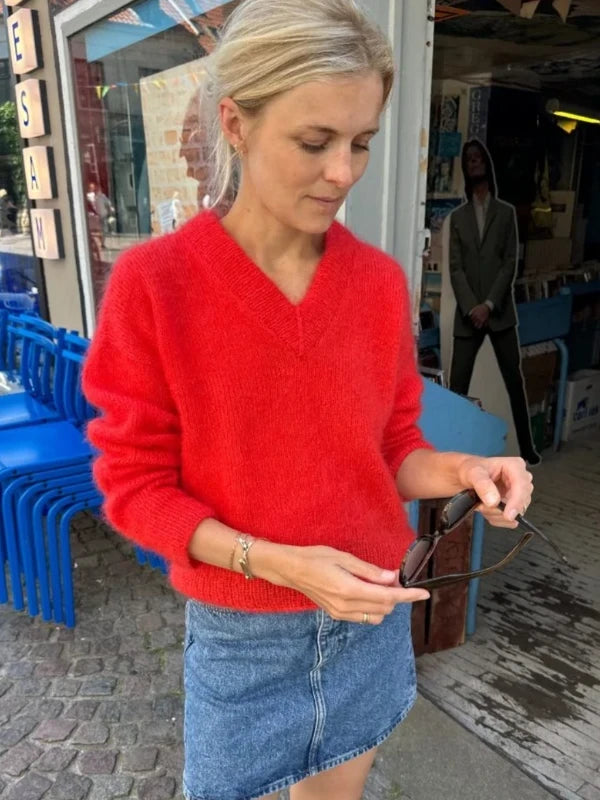 A woman in a vibrant red Petiteknit Stockholm Sweater V-neck neuleohje, Suomi and denim skirt stands outside a shop, looking down while holding sunglasses. Blue stacked chairs and a cardboard cutout are visible in the background.