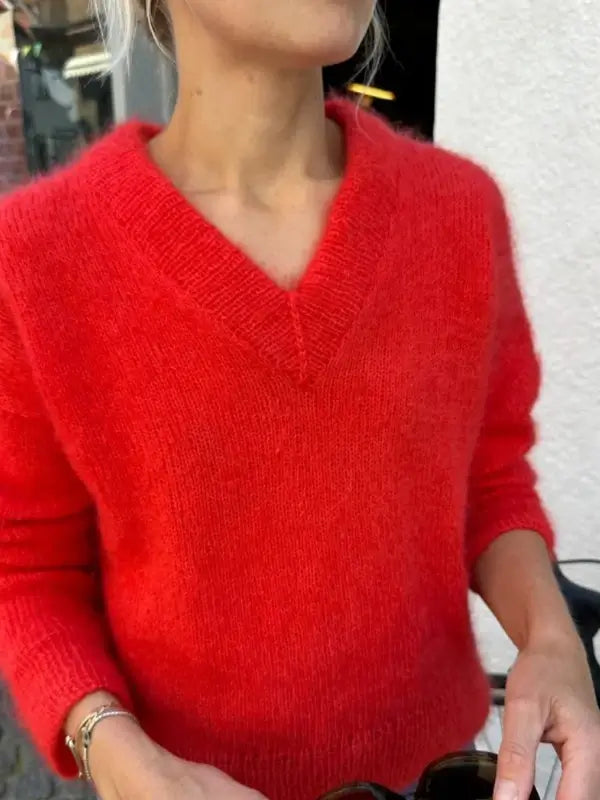 A person wearing a fuzzy, bright red Petiteknit Stockholm Sweater V-neck neuleohje, Suomi, crafted with soft silkkimohair yarn, stands outdoors holding sunglasses. Their head is turned slightly to the side, and their face is mostly out of frame.