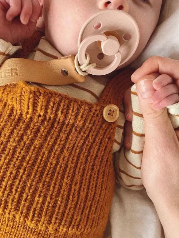 A baby wearing the rust-colored PetiteKnit Willum’s Overalls neuleohje - Suomi and a striped shirt holds an adult’s hand while using a beige pacifier.
