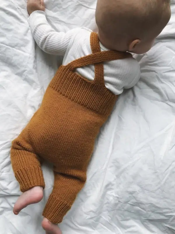 A baby in a white long-sleeve shirt and brown knitted PetiteKnit Willum’s Overalls neuleohje - Suomi lies on a white blanket, facing down with legs stretched out and arms bent—a cozy look perfect for showing off your favorite neuleohje.