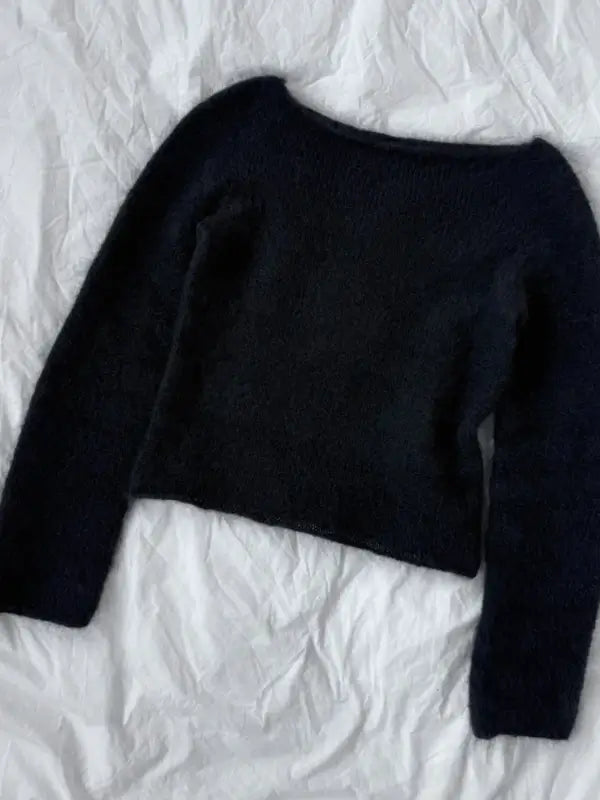 A PetiteKnit Bea Blouse neuleohje sweater with long sleeves in black is laid flat on a white, slightly wrinkled fabric background.
