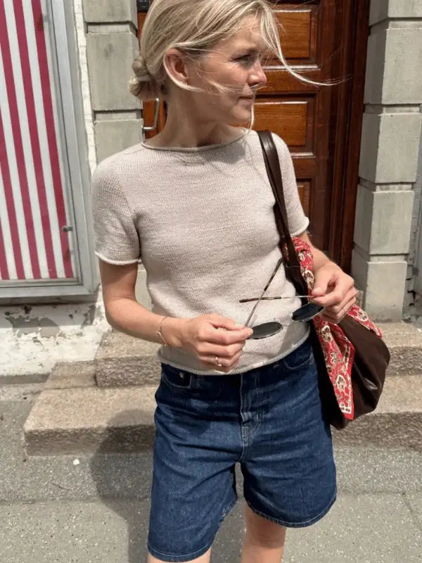 A blonde-haired woman with a low bun stands outdoors holding sunglasses. She wears the beige PetiteKnit Ivy Tee neuleohje, denim shorts, and a patterned tote bag. A striped window and wooden door are in the background.