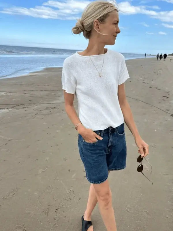 A woman with blonde hair in a low bun walks on a sandy beach wearing the PetiteKnit Cloud tee neuleohje, denim shorts, and black sandals while holding sunglasses. The ocean and a cloudy sky are visible in the background.