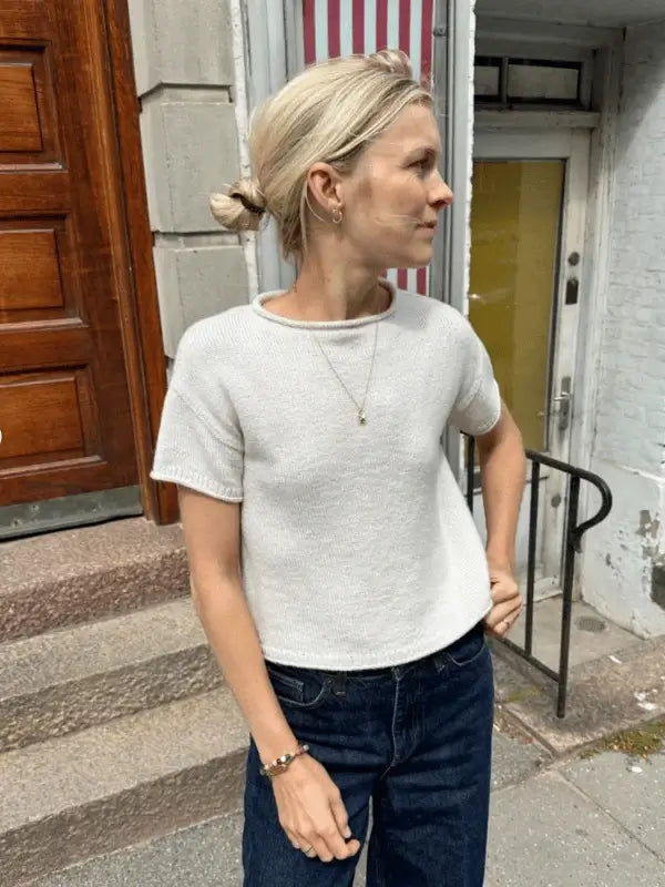 A person with blonde hair in a low bun wears a white PetiteKnit Cloud tee neuleohje short-sleeve sweater, dark jeans, and jewelry, standing outside by stone steps and a wooden door, looking to the side.