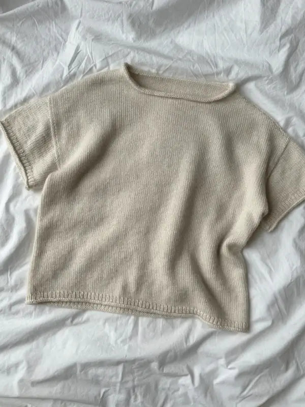 A beige, short-sleeve knit sweater made using the PetiteKnit Cloud tee neuleohje pattern is displayed flat on a wrinkled white fabric background.