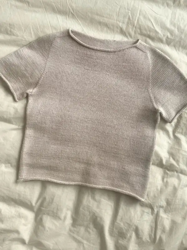 A light gray, short-sleeved knitted top is displayed on a white, slightly wrinkled background. The seamless design and round neckline make it ideal for creating with the PetiteKnit Ivy Tee neuleohje pattern.