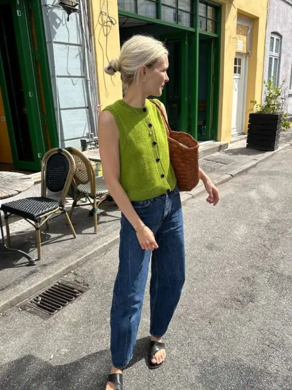 A woman with blond hair in a low bun wears a sleeveless green knit top made with the PetiteKnit Maggie Vest neuleohje, paired with blue jeans, black sandals, and a woven brown bag as she stands on a quiet street near outdoor café chairs.