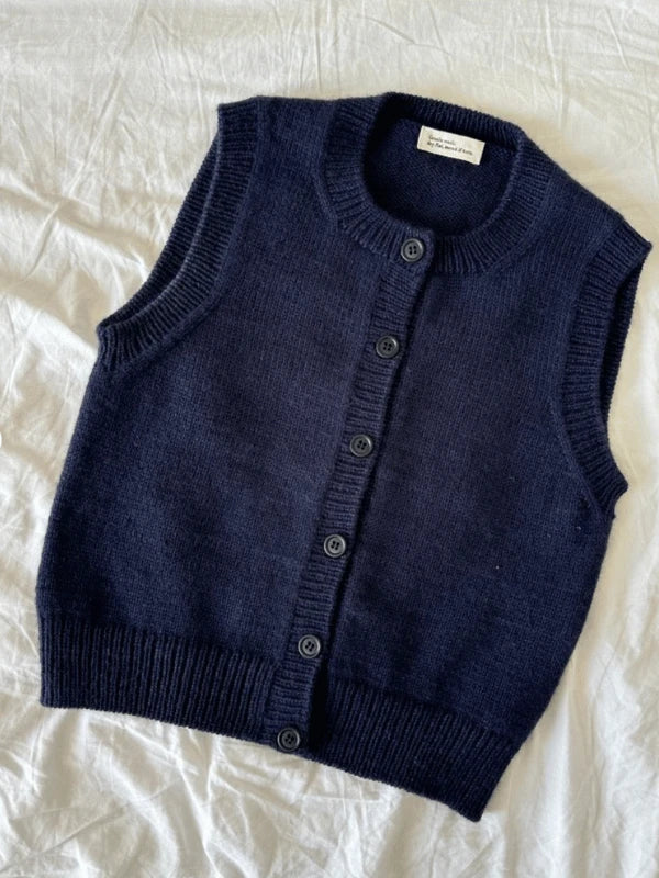 The PetiteKnit Maggie Vest neuleohje is a sleeveless, dark navy blue knit button-up with ribbed hems and a round neckline, shown laid flat on crumpled white fabric—perfect inspiration for your next knitting project.