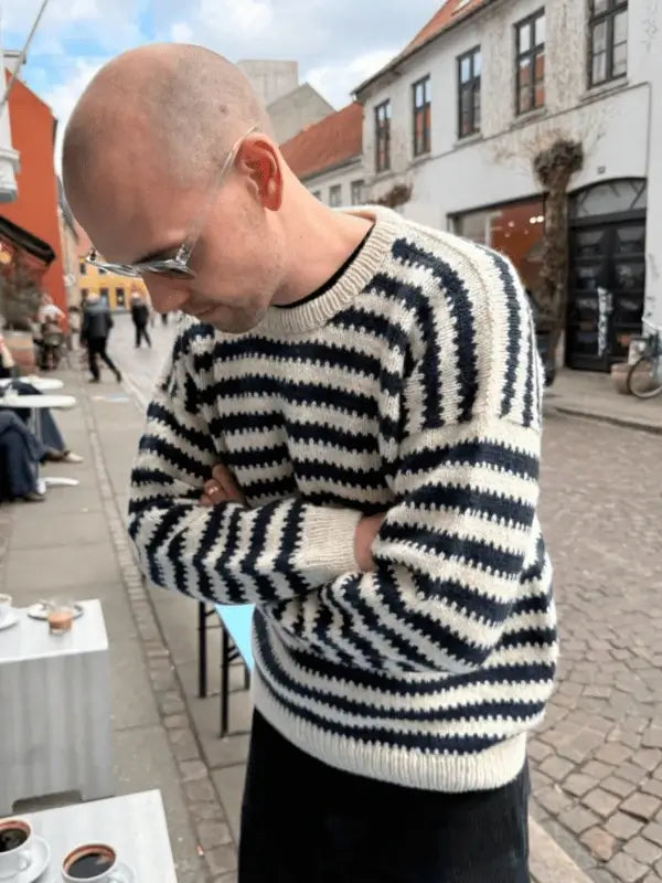 A person dubbed the Otto Sweater Man stands on a cobblestone street wearing glasses and a striped black-and-white PetiteKnit Otto Sweater neuleohje. Cafés and bicycles line the street beneath partly cloudy skies.