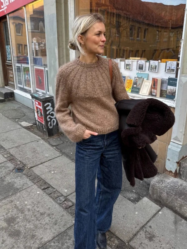 Wearing a PetiteKnit Sunday Sweater neuleohje, Suomi and blue jeans, a woman stands on a city sidewalk holding a dark coat, with postcards and art displayed in a shop window behind her.