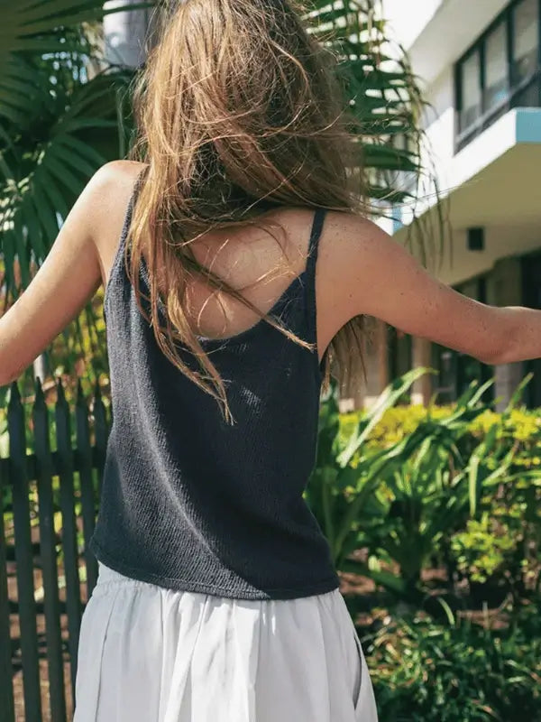 A woman with long hair stands outdoors by greenery and sunlight, wearing a dark sleeveless top made from the Sandnes Garn Clementine Top neuleohje ENG (PDF) and Sandnes Garn Tynn Line, holding knitting needles and yarn.