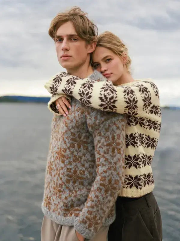 A woman in a cream snowflake neulesetti hugs a man wearing a grey Sandnes Garn Alvar – neulesetti Atlas by a lake.